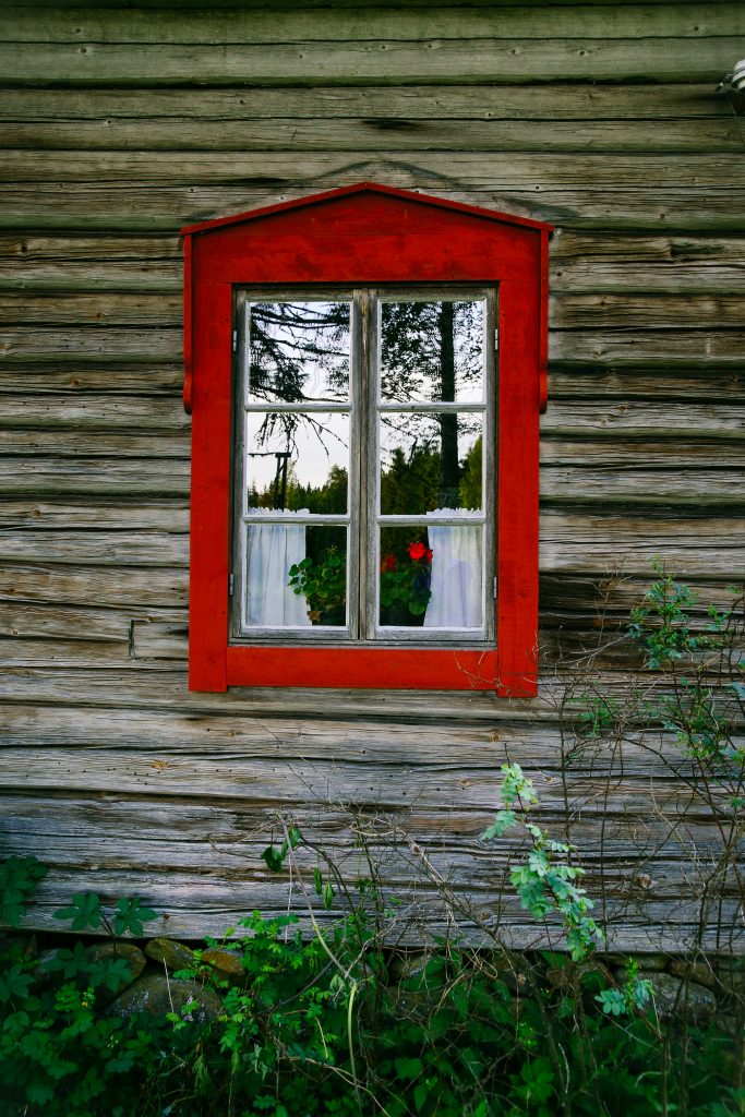 Urjalan museon vanhan hirsirakennuksen idyllinen punareunuksinen ikkuna, jossa on pitsiverhot ja pelargunia ikkunalla.